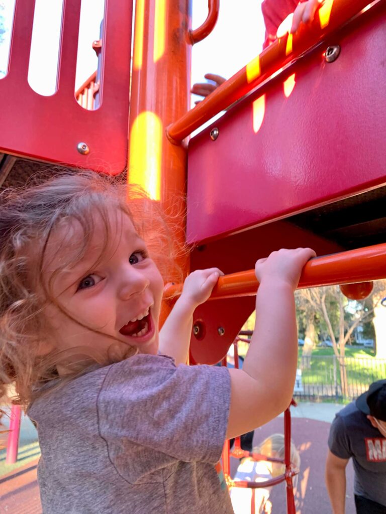little girl looking back grinning as she climbs on a play structure at the park