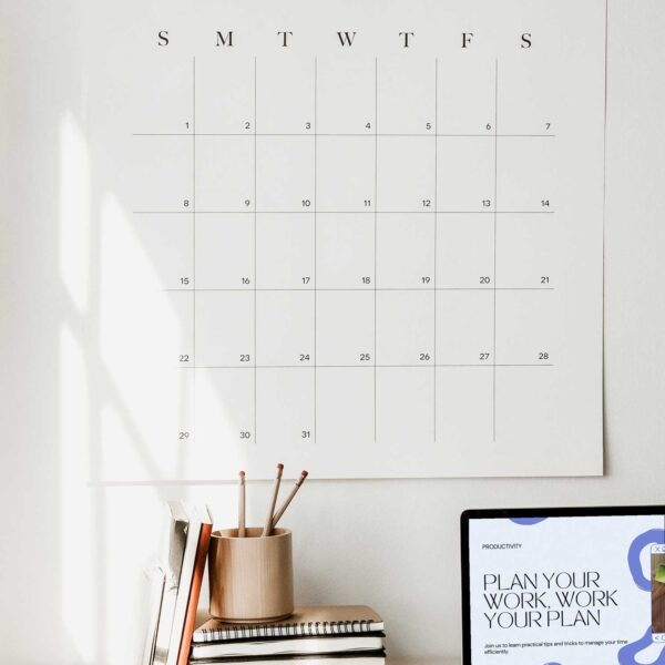 a 30-day wall calendar hanging above a desk with a stack of books and a laptop