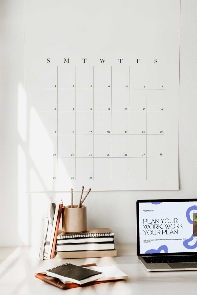 a 30-day wall calendar hanging above a desk with a stack of books and a laptop