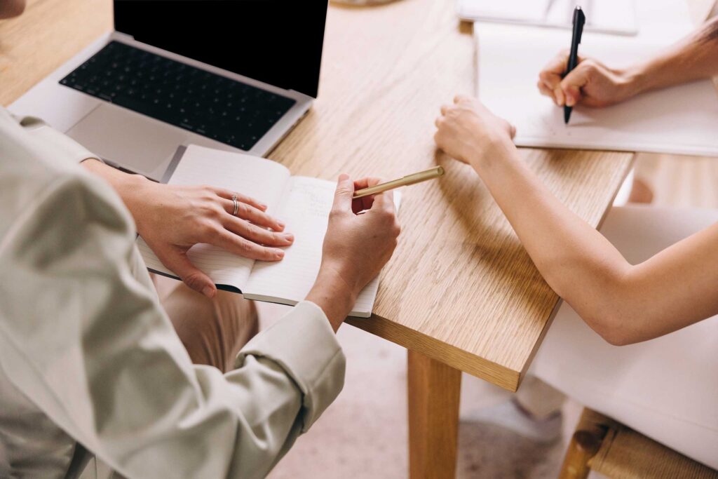 overhead angle of three sets of arms/hands taking notes at a table with a laptop opened