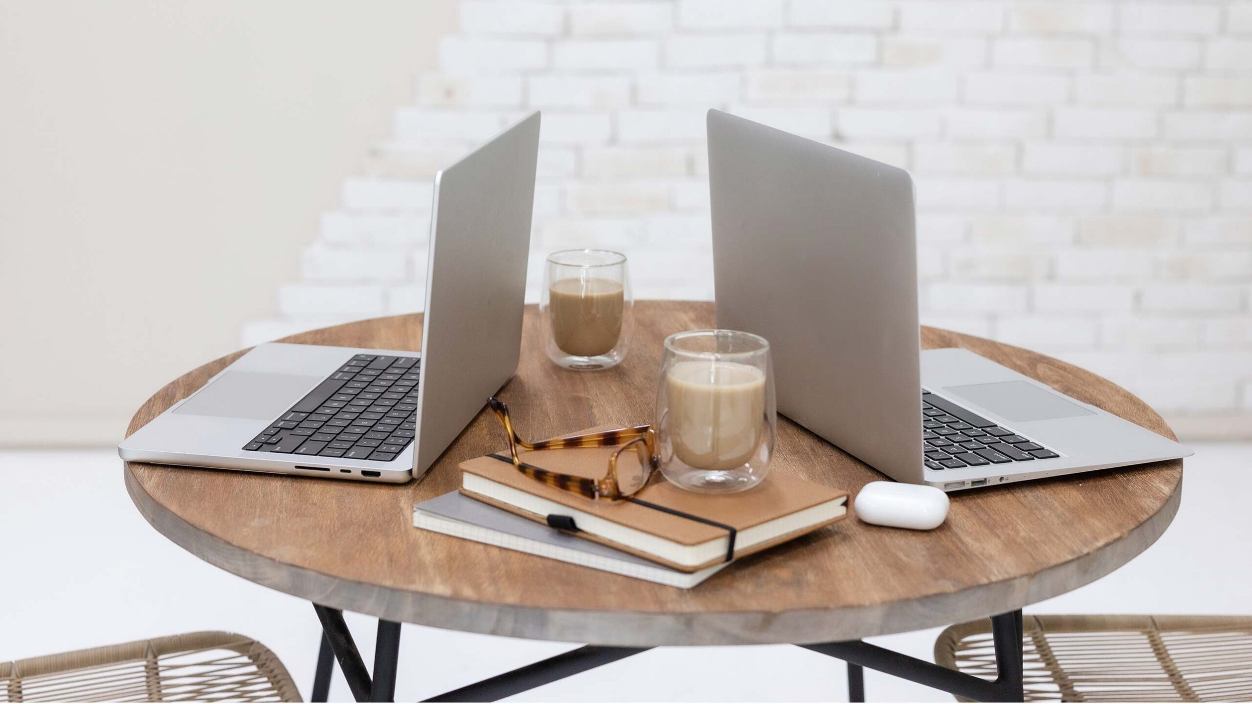 two laptops facing one another on a wooden cafe table with notebooks and glasses stacked beside them